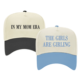 Image of two baseball caps. The left cap has a cream top with a black bill and features the phrase 'IN MY MOM ERA' embroidered in black. The right cap has a cream top with a light blue bill and displays the embroidered phrase 'THE GIRLS ARE GIRLING' in blue